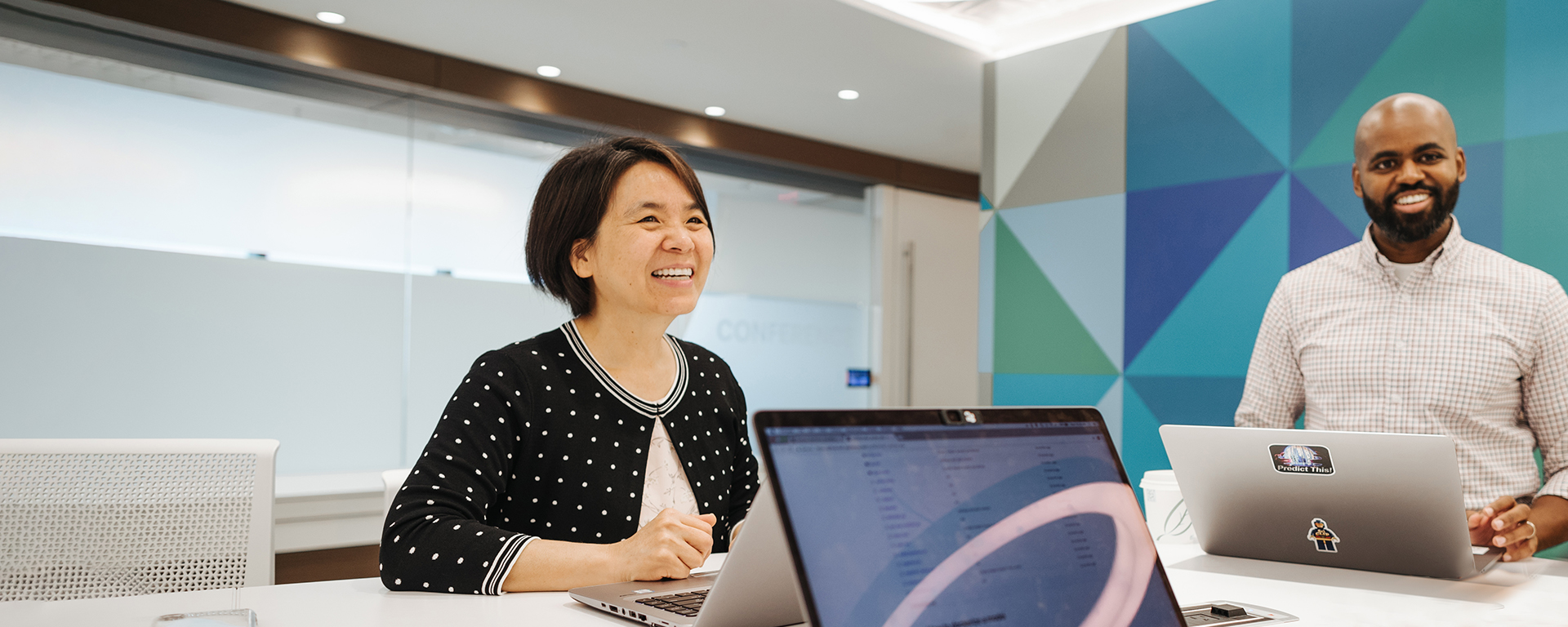 Capital One Tech associates stand at a conference room table and collaborate on their laptops