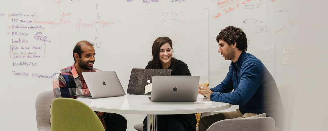 Three Capital One associates sit around a table with laptops with white board writings on the wall behind them