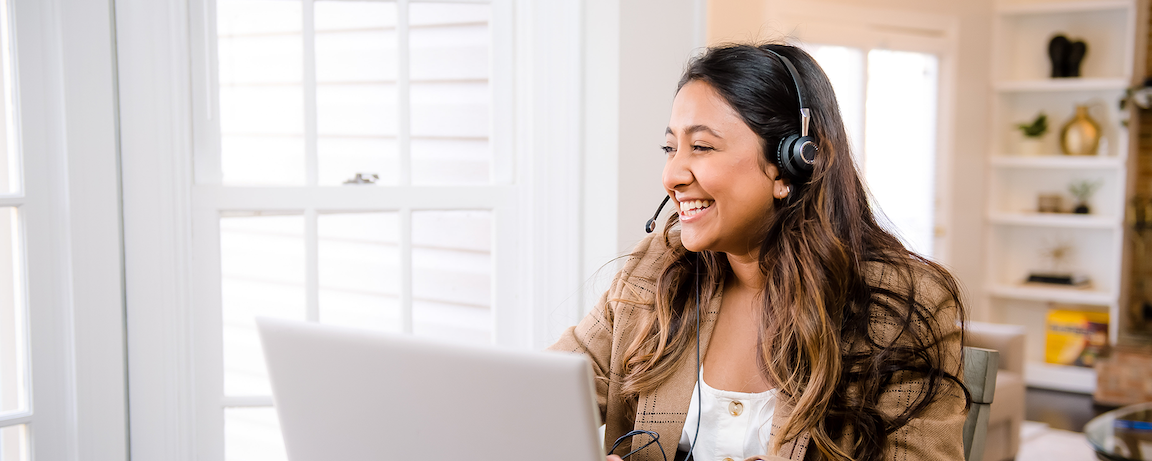 Woman smiles wearing a pair of headphones and typing on a silver laptop.