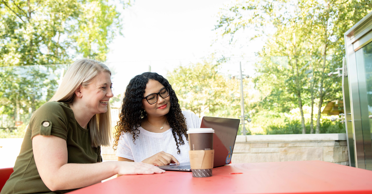 Two Capital One associates sit outside at a red table and look at a laptop