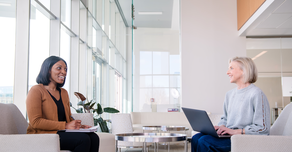 Two Capital One associates sit on couch chairs with their laptops and talk