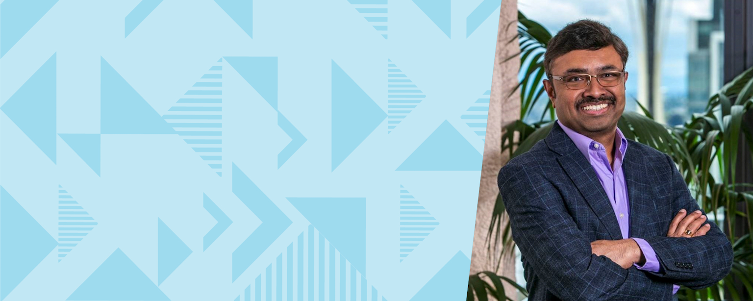 Capital One leader Prem stands in front of a tree in a suit smiling with his arms crossed