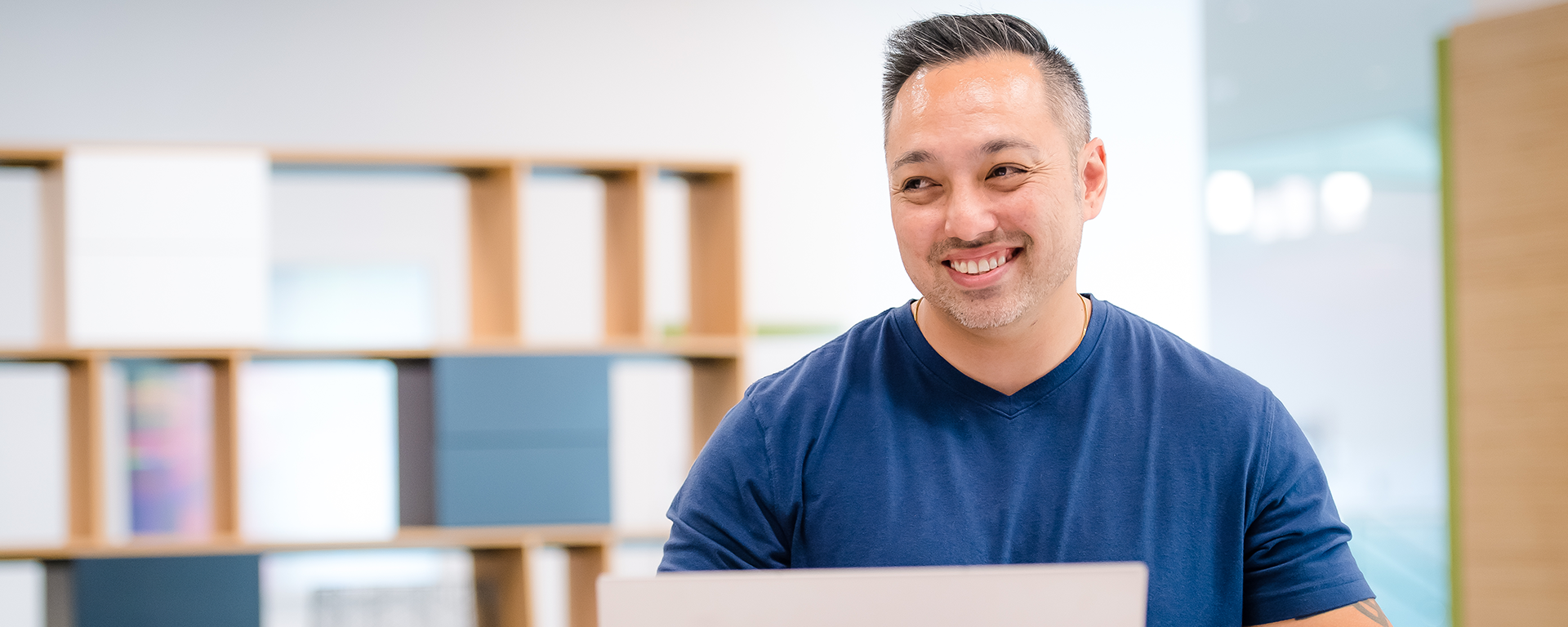 A man smiles in a blue t-shirt while holding a cup of coffee and looking at a computer